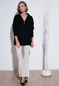 Un pull noir surdimensionné avec un col zippé et des poches décorées de clous, associé à un pantalon évasé beige clair et des talons noirs.
