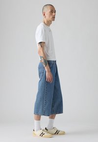 Camiseta blanca de manga corta, pantalones culotte de mezclilla holgados y zapatillas crema con rayas marrones. Presenta tatuajes visibles en el brazo. Fondo neutro.