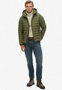 Oliwkowa kurtka puchowa z kapturem, biała sweter z golfem o prążkowanej fakturze, niebieskie dżinsy i brązowe botki sznurowane. Posiada pikowaną strukturę i zamek błyskawiczny.