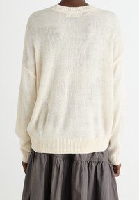 Ljusbeige stickad tröja med löst snitt, ribbad nederkant och sänkta axlar, i kombination med en grå kjol. Texturerat, andningsbart material.