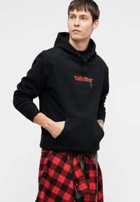 Μαύρη κουκούλα με εμπρόσθια τσέπη και κόκκινο λογότυπο "Santa Cruz"; σε συνδυασμό με ένα κόκκινο και μαύρο καρό πουκάμισο δεμένο γύρω από τη μέση.