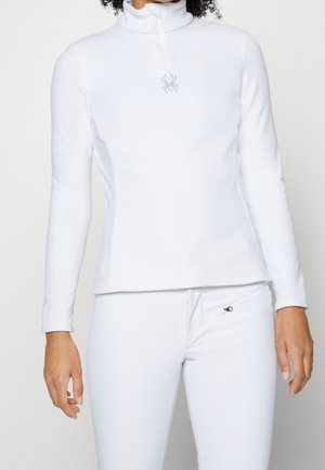 Kvinde iført hvid langærmet lynlås-top med edderkop-logo og matchende hvide bukser med lynlåslomme.