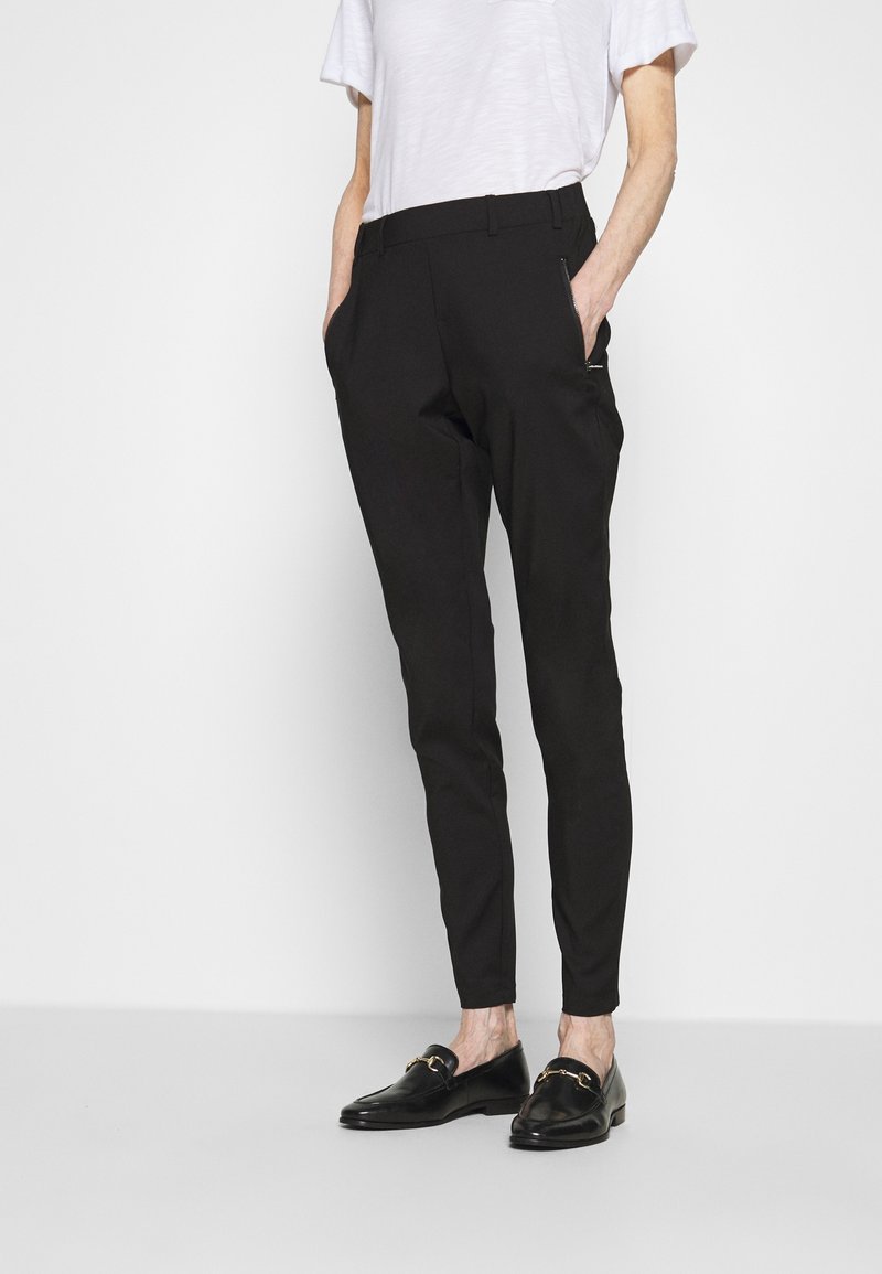 Homme portant un pantalon noir slim avec des poches zippées, une chemise blanche à manches courtes et des mocassins noirs avec un détail chaîne dorée, debout les mains dans les poches.