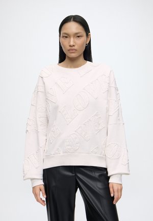 Femme aux longs cheveux noirs porte un sweat-shirt blanc oversize embossé du texte « LOVE » et un pantalon en cuir noir sur fond blanc uni.