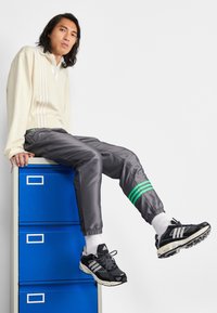 Ljus beige polo med vertikala vita ränder, grå träningsbyxor med gröna detaljer, svarta sneakers, blå arkivskåp i bakgrunden.