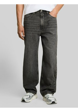 Mann mit locker sitzenden dunkelgrauen Jeans und weißen Sneakers, mit einer Hand in der Tasche vor einfarbigem Hintergrund.