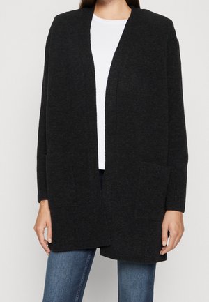 Vrouw draagt een lange, open vallende zwarte cardigan met twee zakken aan de voorkant, over een wit shirt en een blauwe spijkerbroek.