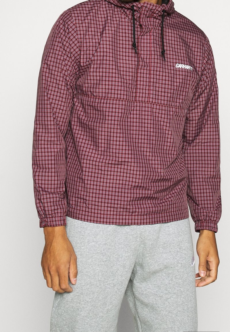 Rode en geruite lichtgewicht jas met capuchon; elastische manchetten; voorvak; voorzien van het "Carhartt" logo in het wit. Gepaard met grijze trainingsbroeken.