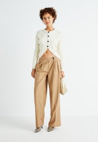 Cardigan court en maille torsadée crème avec des boutons noirs, associé à un pantalon beige à taille haute et plis, et des talons aiguilles métalliques.