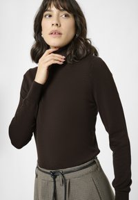 Mujer con cabello oscuro hasta los hombros, vistiendo un suéter de cuello alto marrón y pantalones de cintura alta a cuadros con un cinturón negro, con la mano cerca de la barbilla.
