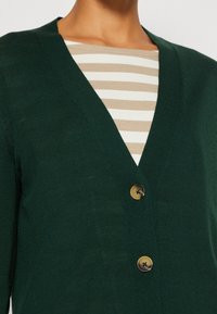 Cárdigan verde con diseño de escote en V, hecho de tela de punto. Presenta dos botones marrones y está superpuesto a una camisa a rayas beige y blanca.