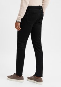 Jeans slim fit noirs avec une texture lisse, dotés de deux poches arrière et de détails de couture subtils, associés à des chaussures grises.