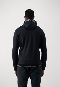 Sweat à capuche noir avec une capuche discrète et des poignets côtelés, arborant le logo PeakPerformance sur le col et l'ourlet inférieur. Texture de tissu lisse.