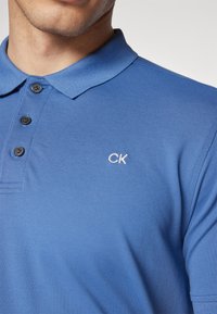 Polo de manga curta azul feito de algodão texturizado, com uma pala de três botões e um pequeno logotipo branco da CK no peito.