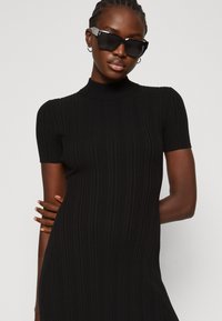 Black ribbed dress with a high neck, short sleeves, and a form-fitting silhouette. Model wears bold black sunglasses, emphasizing modern design.