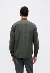 Camisa de manga larga en tejido verde oscuro, con cuello abotonado, yugo trasero y dobladillo curvado. Diseño minimalista sin patrones visibles.