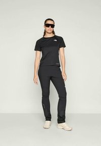 Svart atletisk outfit med kortärmad topp och åtsittande byxor, med logotypdetaljer. Modellen bär solglasögon. Slät, elastisk tyg.