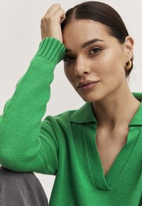 Zeleni pleteni pulover s polo ovratnikom i rebrastim manžetama. Model ima neutralan make-up i zlatne naušnice na obruče.