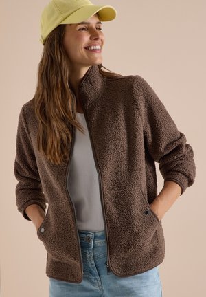 Femme souriante portant une veste polaire marron zippée, une chemise blanche, un jean bleu clair et une casquette de baseball jaune, les mains dans les poches de la veste.