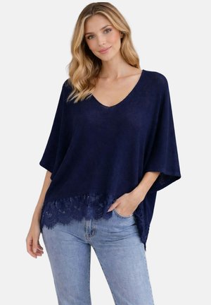 Femme portant un haut ample bleu marine à col en V avec bordure en dentelle et un jean bleu clair, posant avec une main dans la poche sur fond blanc.