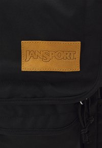 Juodas audinio kuprinė su odos panašiu rudu pleistru, ant kurio yra "JANSPORT" logotipas. Yra reguliuojamos diržai ir priekinis kišenė.