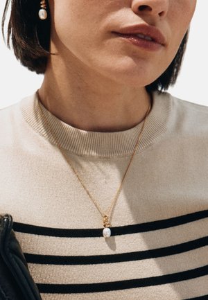 Kvinde iført beige trøje med sorte striber, guld kædehalskæde med perlevedhæng og guld dråbeørering med perle.