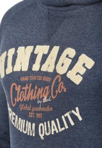 Dunkelblaues Sweatshirt mit strukturierter Oberfläche, das ein großes Grafikdesign im Vintage-Stil in Creme und Orange zeigt, mit den Worten "Vintage Clothing Co." und "Premium Quality."