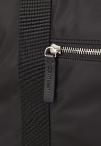 Zwarte stoffen tas met een gestructureerde schouderband, zilveren rits en een zwart leren label met de opdruk "Calvin Klein". Minimalistisch ontwerp met functionele details.