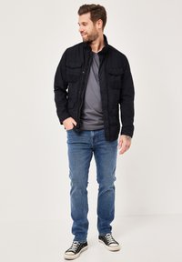 Schwarze Jacke mit vier Fronttaschen, graues T-Shirt, blaue Jeans und schwarze Turnschuhe. Das Model steht in einer entspannten Pose.