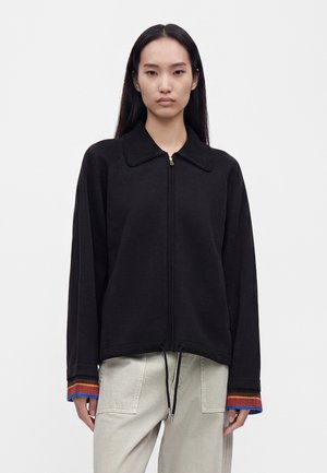 Jeune femme aux longs cheveux noirs porte une veste noire zippée avec un col côtelé et des poignets rayés, ainsi qu’un pantalon beige clair avec des poches avant.