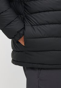 Schwarze Steppjacke mit gesteppten Partien, einem Stehkragen und seitlichen Taschen. Das Material wirkt glatt und isoliert.