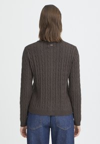 Kobieta o brązowych włosach do ramion, ubrana w ciemnobrązowy sweter z grubym splotem i niebieskie dżinsy, stoi tyłem na jednolitym tle.