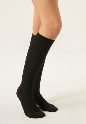 Gambe con calzini neri a coste fino al ginocchio su uno sfondo chiaro uniforme, piedi leggermente divaricati con un tallone leggermente sollevato.