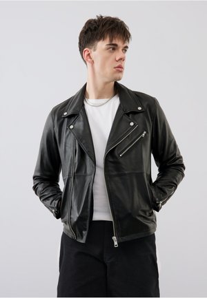 Junger Mann in schwarzer Lederjacke, weißem T-Shirt und schwarzer Hose, steht mit den Händen in den Taschen und schaut seitlich vor einfachem Hintergrund.