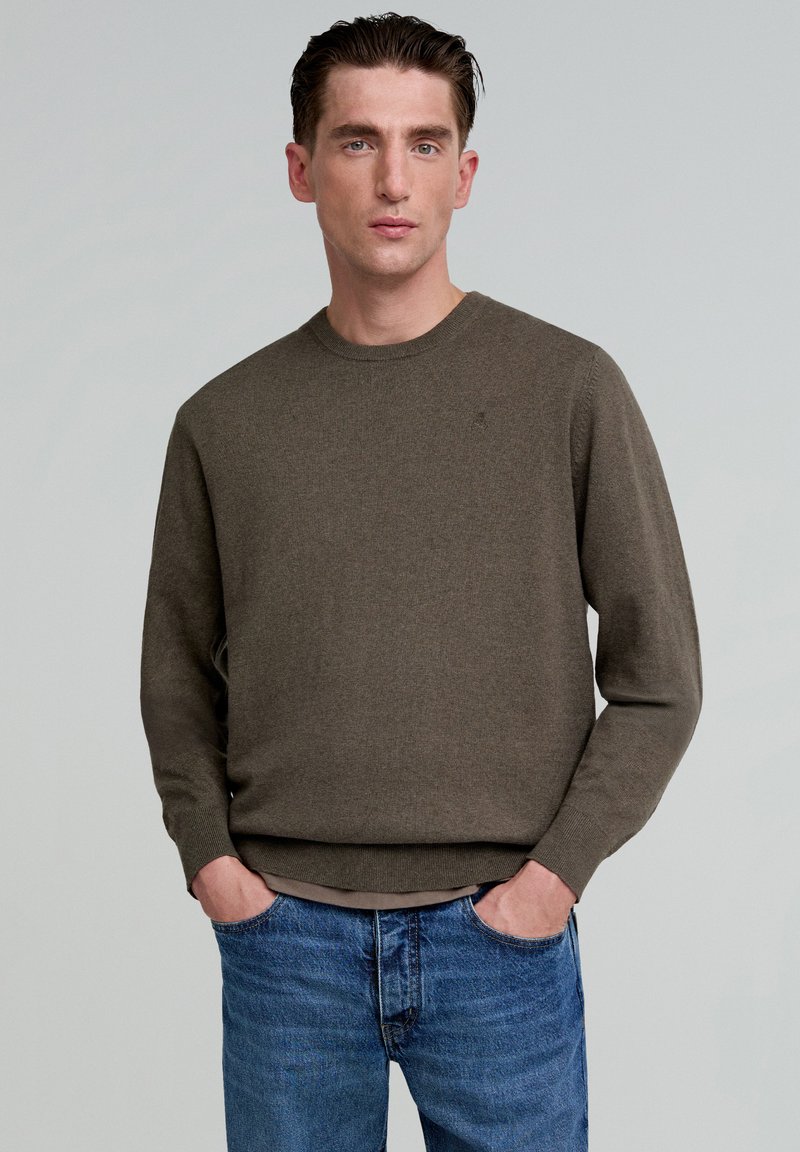 Bruine trui met lange mouwen en ronde hals, gemaakt van een zachte gebreide stof. Het model draagt het over blauwe jeans, wat een ontspannen pasvorm laat zien.