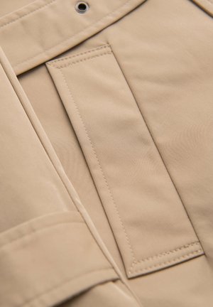 Beige stof met gedetailleerde stiksels en een metalen oogje, close-up van een zak en de omliggende naad op een kledingstuk.