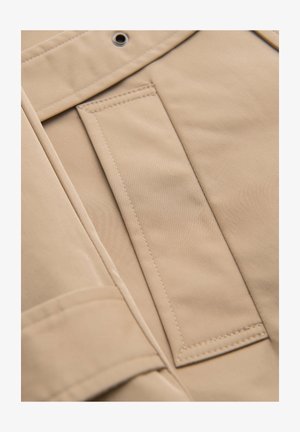 Beige stof met gedetailleerde stiksels en een metalen oogje, close-up van een zak en de omliggende naad op een kledingstuk.