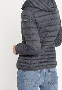 Chaqueta acolchada gris con capucha, con diseño acolchado y logotipo de CMP en la manga. Se lleva con vaqueros azules, mostrando una estructura ajustada.