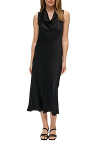 Czarna sukienka bez rękawów z drapowanym dekoltem, gładkiego satynowego materiału i rozkloszowanym dołem, zestawiona z czarnymi sandałami na cienkich paskach.