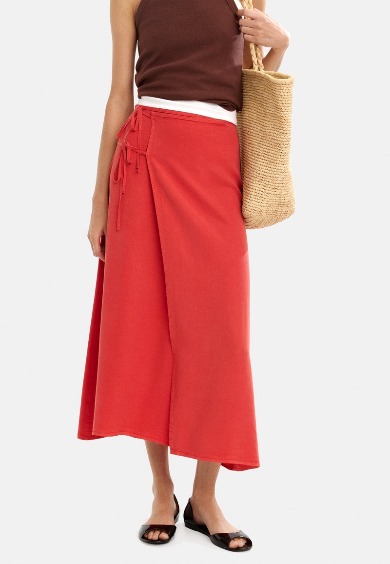 Czerwona spódnica o kroju wrap, wykonana z miękkiego materiału, z bocznym wiązaniem i długością do łydki. Połączona z brązowym topem na ramiączkach i sandałami.