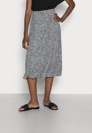 Frau trägt ein schwarzes Oberteil, einen schwarz-weiß gepunkteten Midi-Rock und schwarze flache Sandalen, steht mit einer Hand in der Hüfte.