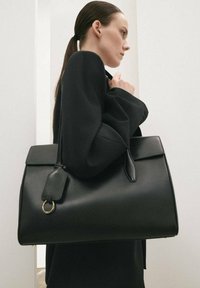 Bolso de cuero negro con un diseño estructurado, que cuenta con un asa plana, un aro para llavero y una textura suave. Minimalista y espacioso.