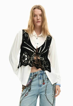 Nuori nainen pukeutuneena valkoiseen nappipaitaan, mustaan virkattuun liiviin ja vaaleansinisiin farkkuihin, joissa on ketju- ja charmiprintti.