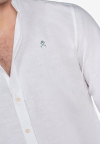 Camisa de lino blanca con un cuello relajado y un pequeño diseño bordado en verde en el pecho. Presenta botones de color claro.