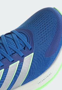 Plava sportska cipela s mrežastim i sintetičkim gornjim dijelom, sadrži srebrne pruge i zelene akcentne detalje. Uključuje vezanje uzicom i teksturiranu potplat.