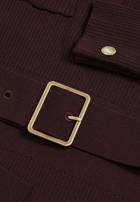 Burgundský ribovaný textil s odpovídajícím páskem se zlatým obdélníkovým sponou a detaily knoflíků na látce.