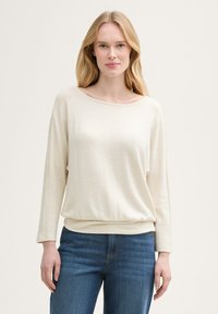 Blondide juustega naine, kes kannab kreemikat pikakäist-sweaterit ja siniseid teksaseid, seisab ühevärvilise heleda tausta ees.