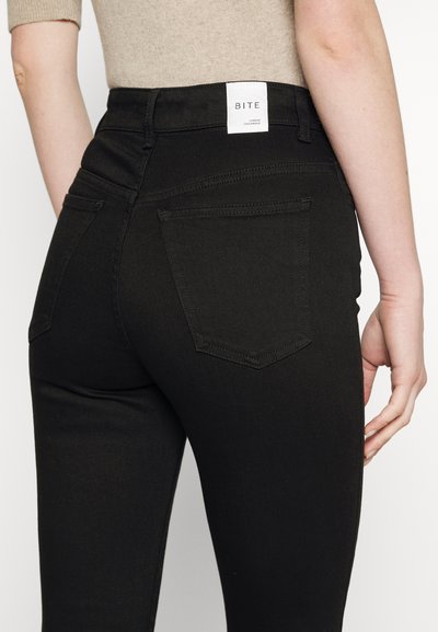 Jeans de denim preto com uma cintura alta, apresentando uma etiqueta branca com a inscrição "BITE" nas costas, dois bolsos e um design ajustado.