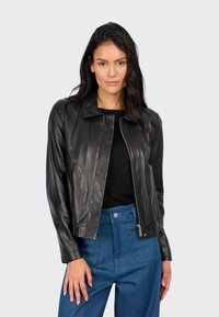 Černá kožená bunda se střihem do rovna, stojáčkem a předním zipem. Nositá přes černou košili, doplněná o kalhoty s vysokým pasem v modré barvě.