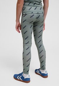 Tight-fitting, olivgröna leggings med ett svart upprepande "HUMMEL" logomönster. Bärs med blå träningsskor med vita detaljer.